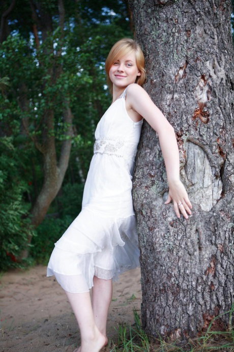 Pretty teen girl removes a white dress to get naked in a sunlit forest area