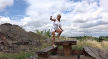 Blonde Lilith in a white dress shows pink while pissing off a windy cliff