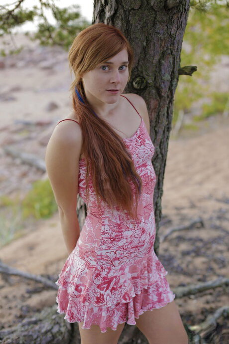Young redhead strips naked in the outdoors against a tree on sandy ground