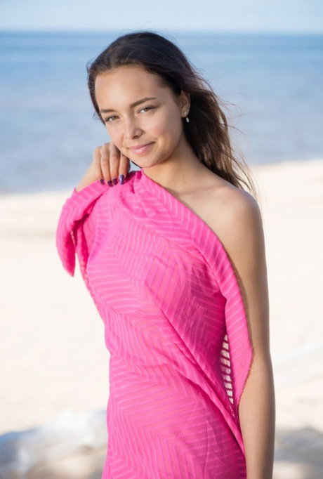 Sweet brunette teen Slava A holds pink fabric while posing nude on a beach